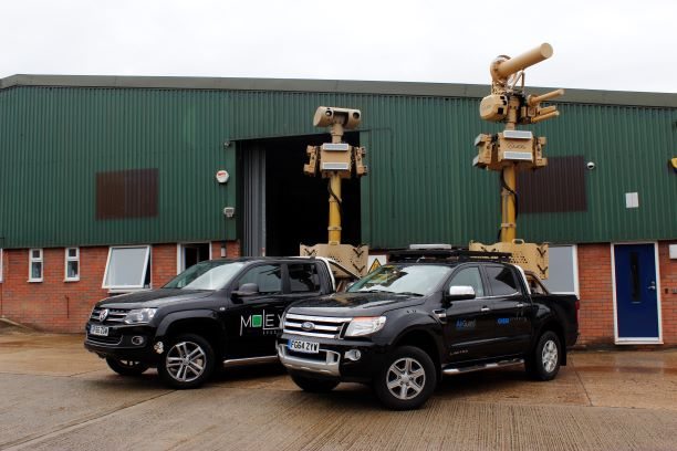 Two pickup trucks carrying mast-mounted surveillance and counter-drone equipment outside a building.