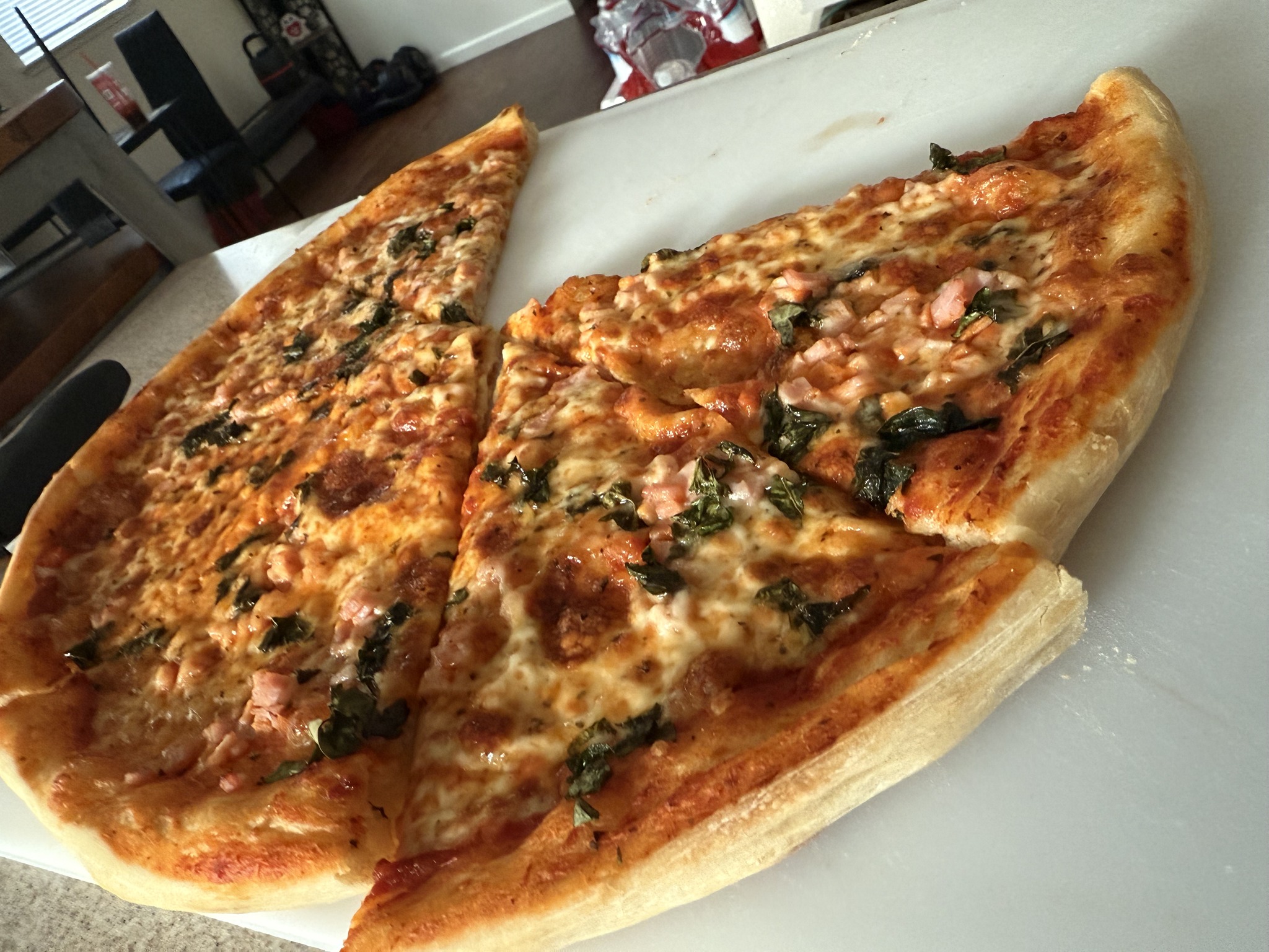 Sliced pizza with browned cheese, pink diced pieces and leafy green toppings on a white cutting board.