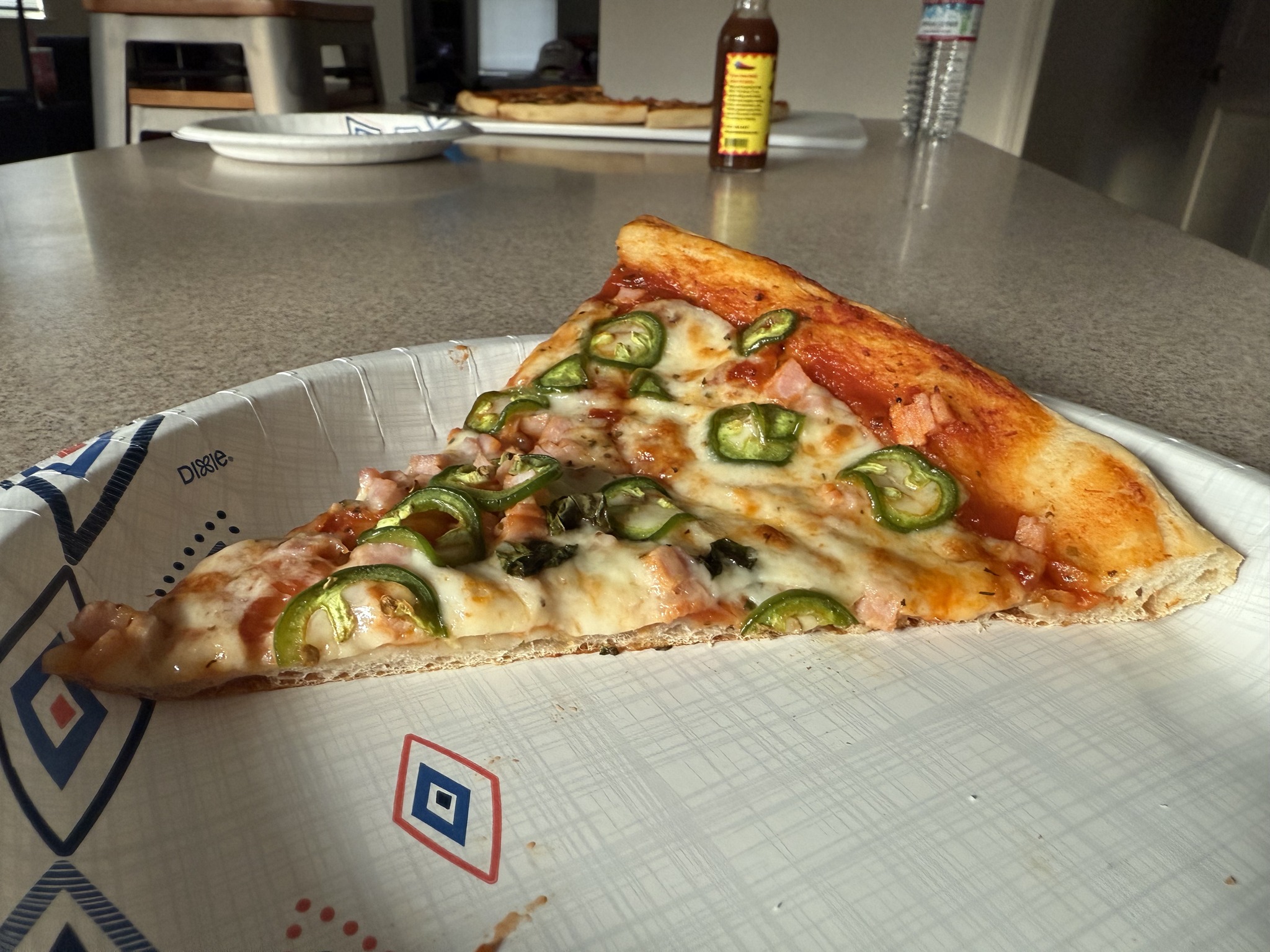 A pizza slice topped with green chili rings, cheese and pink diced pieces on a paper plate.