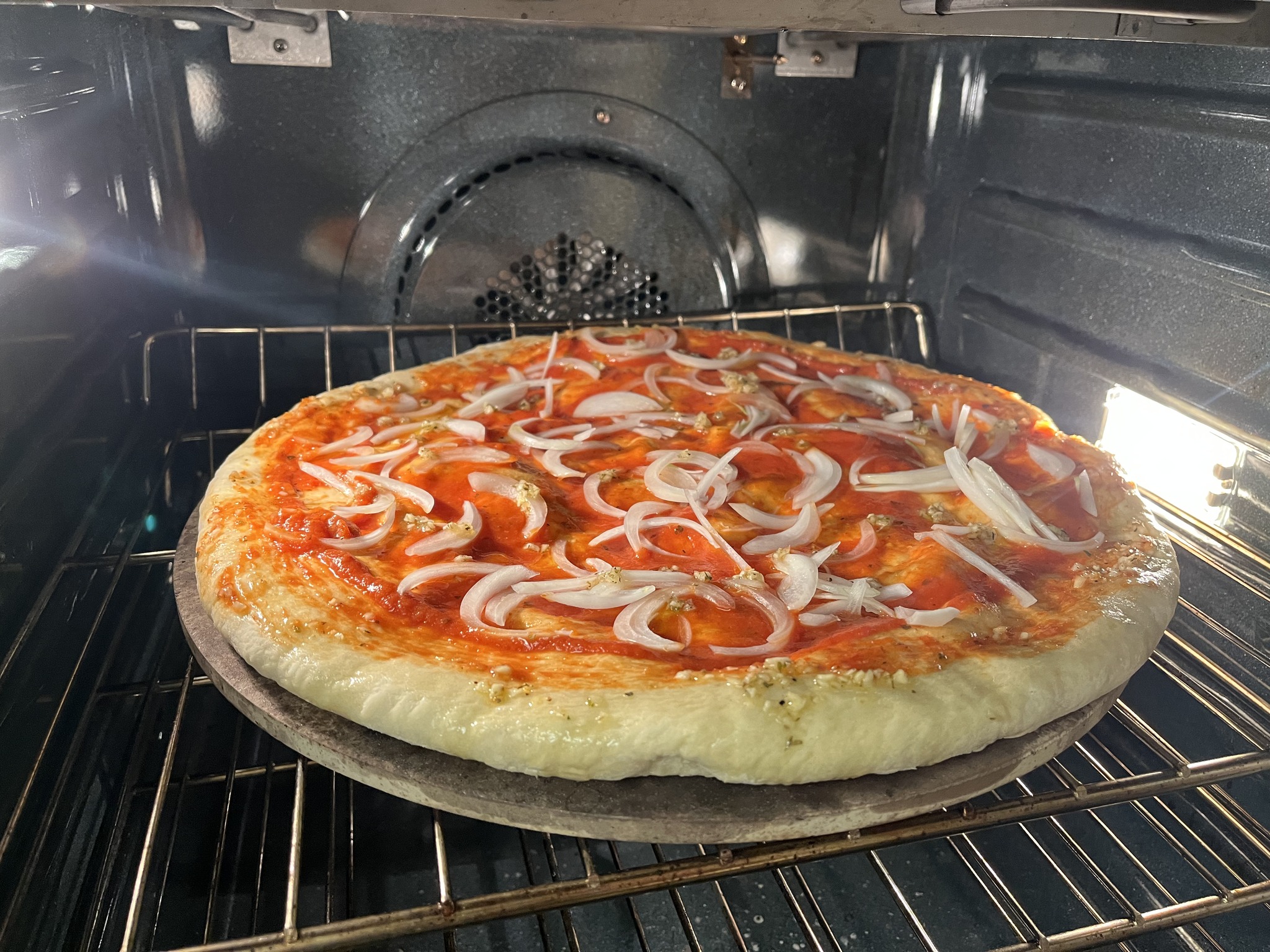 A pizza with red sauce and sliced onion on a round baking stone inside a kitchen oven.