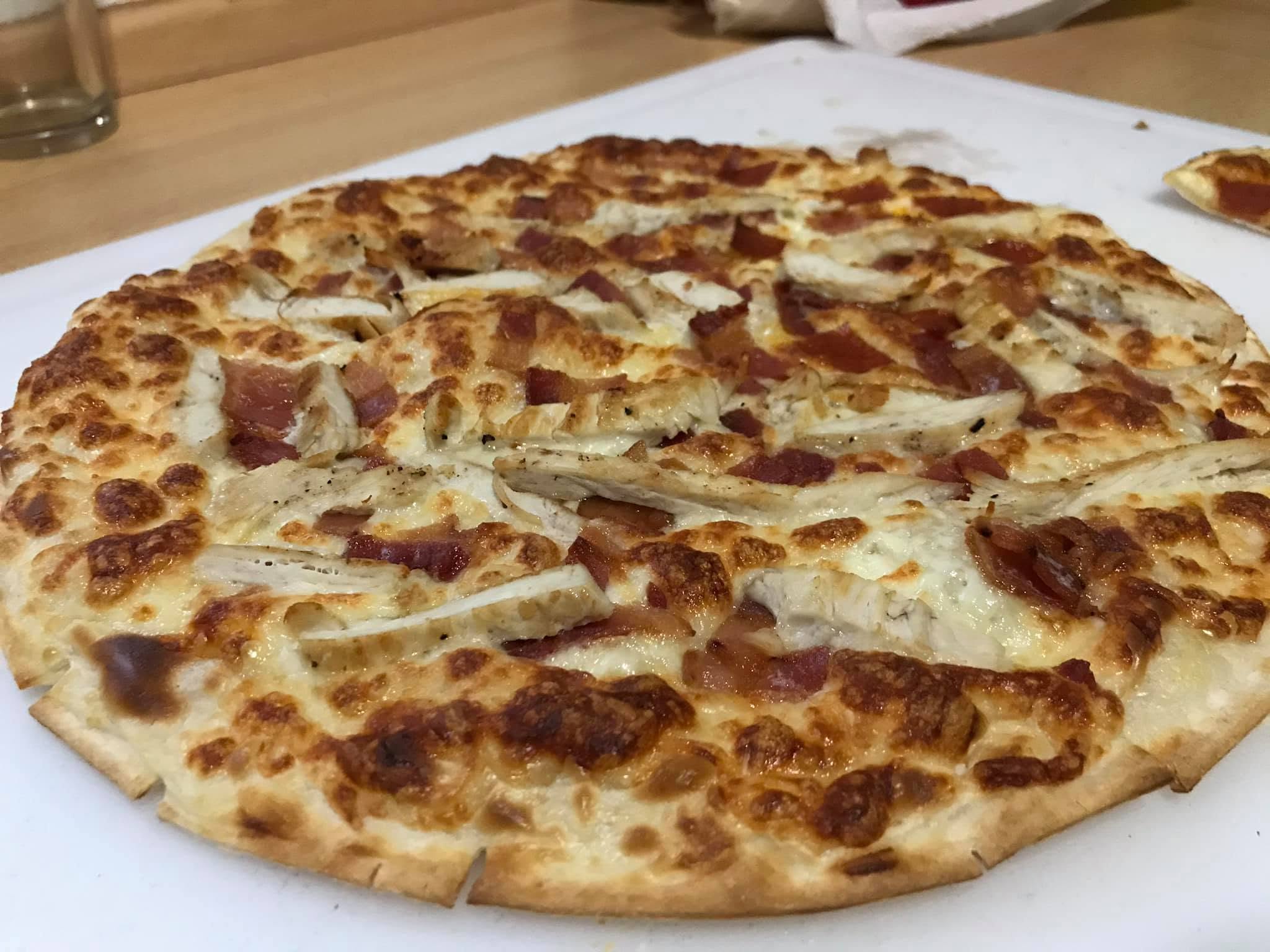 Finished thin-crust flammkuchen topped with browned cheese, chicken strips and pieces of meat, cut into slices on a board.