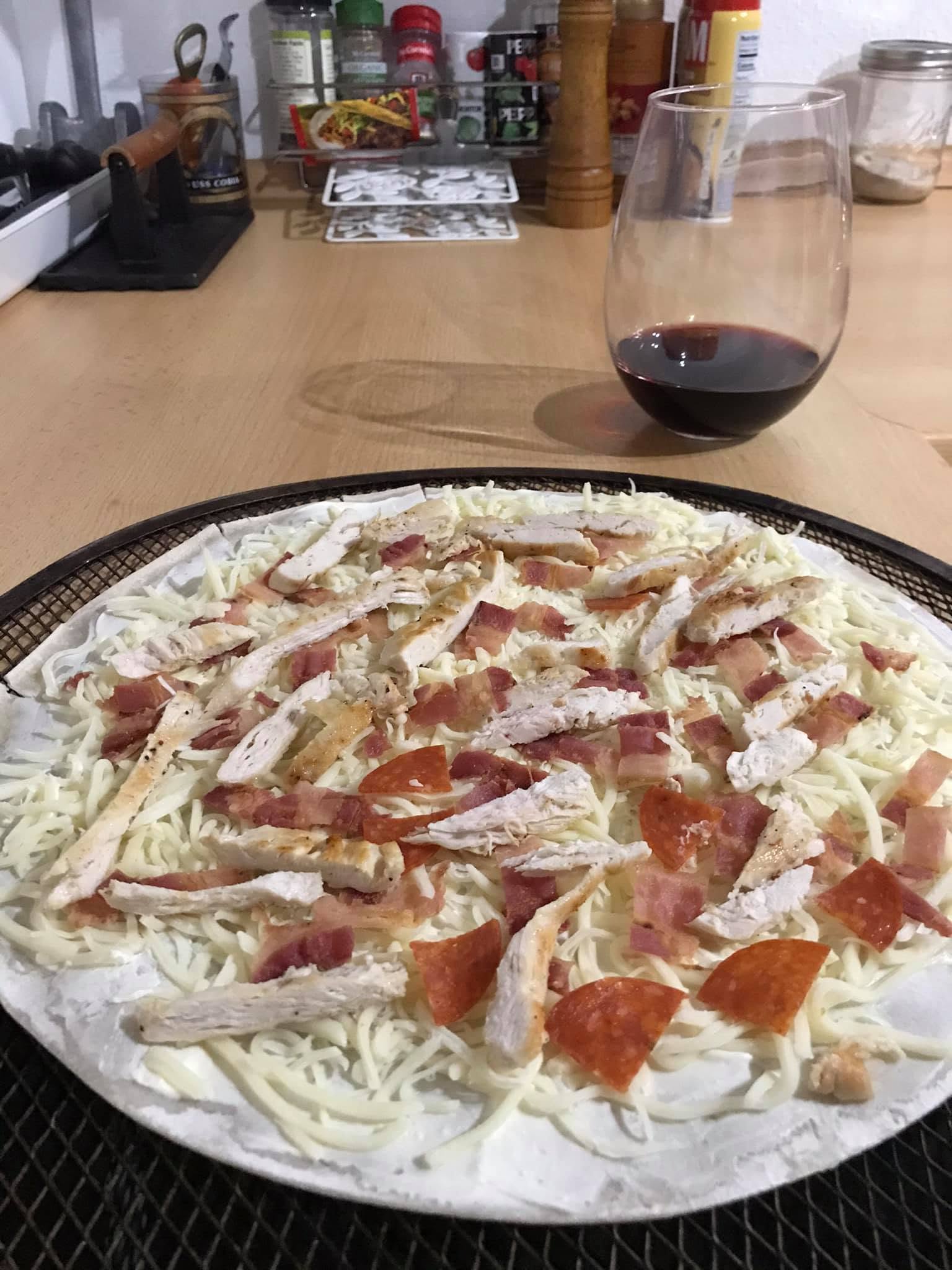 Unbaked flammkuchen with shredded cheese, chicken strips and pieces of bacon, ham and pepperoni on a round mesh baking tray.