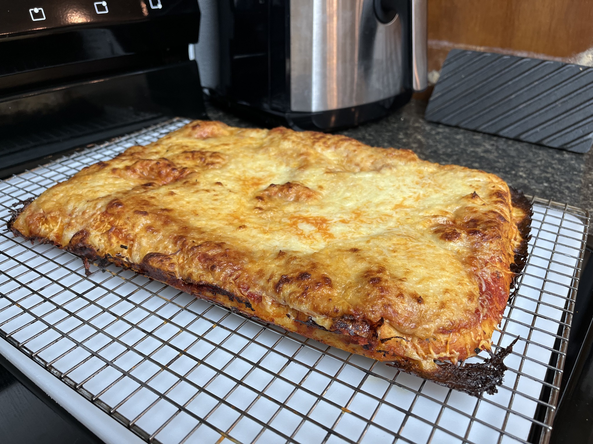 A rectangular cheese pizza with dark, lacy baked-cheese edges resting on a cooling rack.