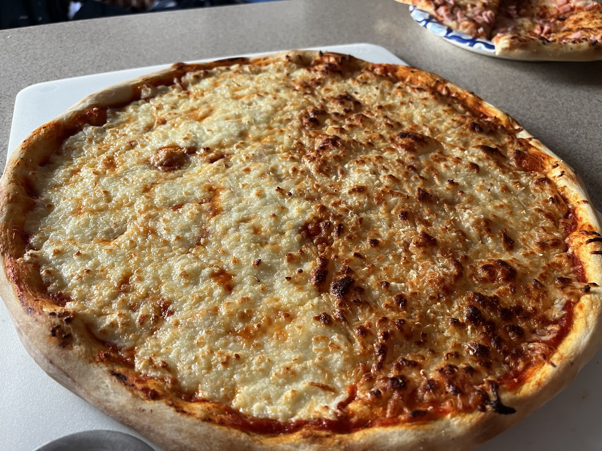A large cheese pizza with pale and deeply browned areas on a white board, with other slices behind it.