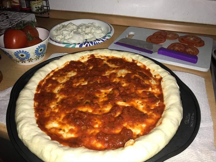 Unbaked pizza dough with a raised edge and red sauce in a round pan, beside chopped cheese and sliced tomatoes.