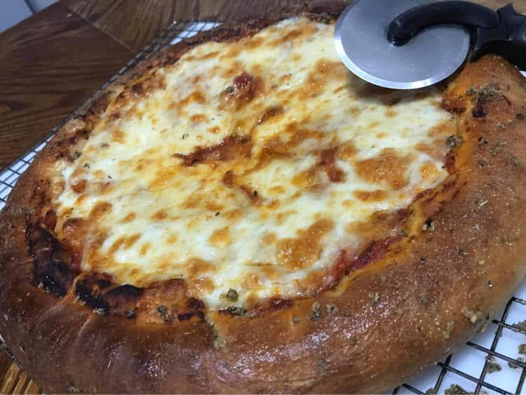 A cheese pizza with a tall crust on a wire cooling rack, with a pizza wheel at the top edge.