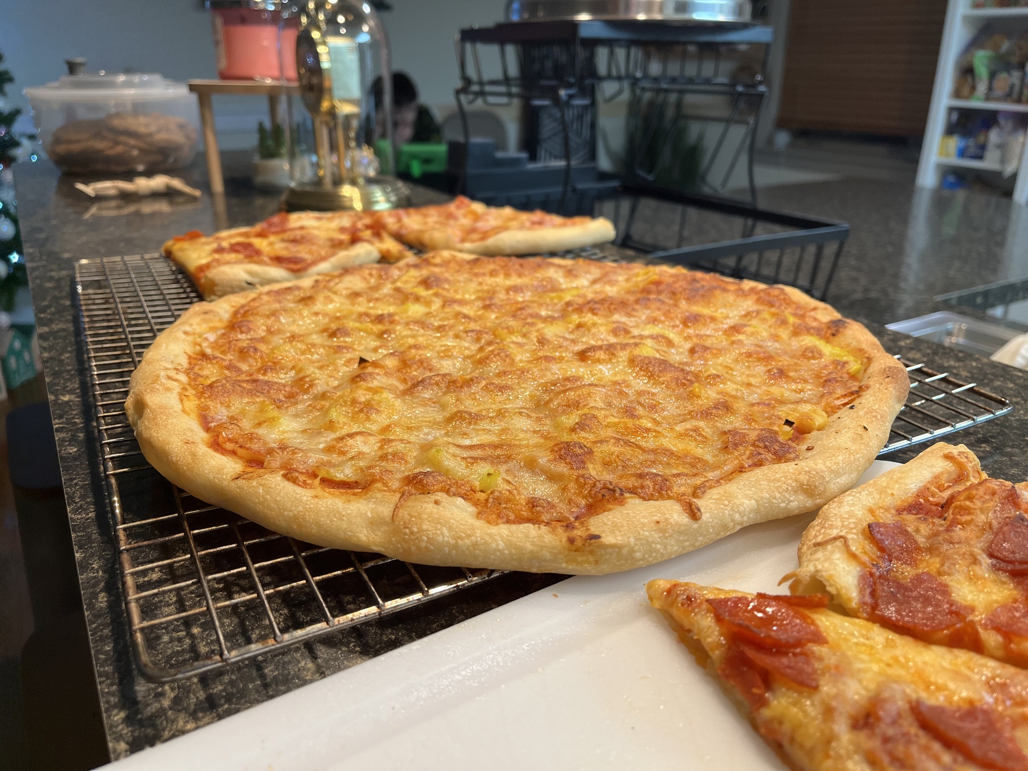 A whole cheese-covered pizza on a cooling rack with sliced pepperoni pizza nearby.