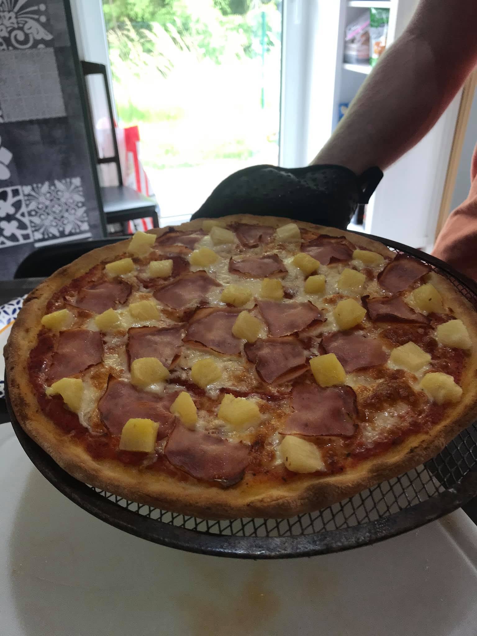 Pizza topped with pineapple chunks and slices that appear to be ham, held on a mesh screen with an oven glove.