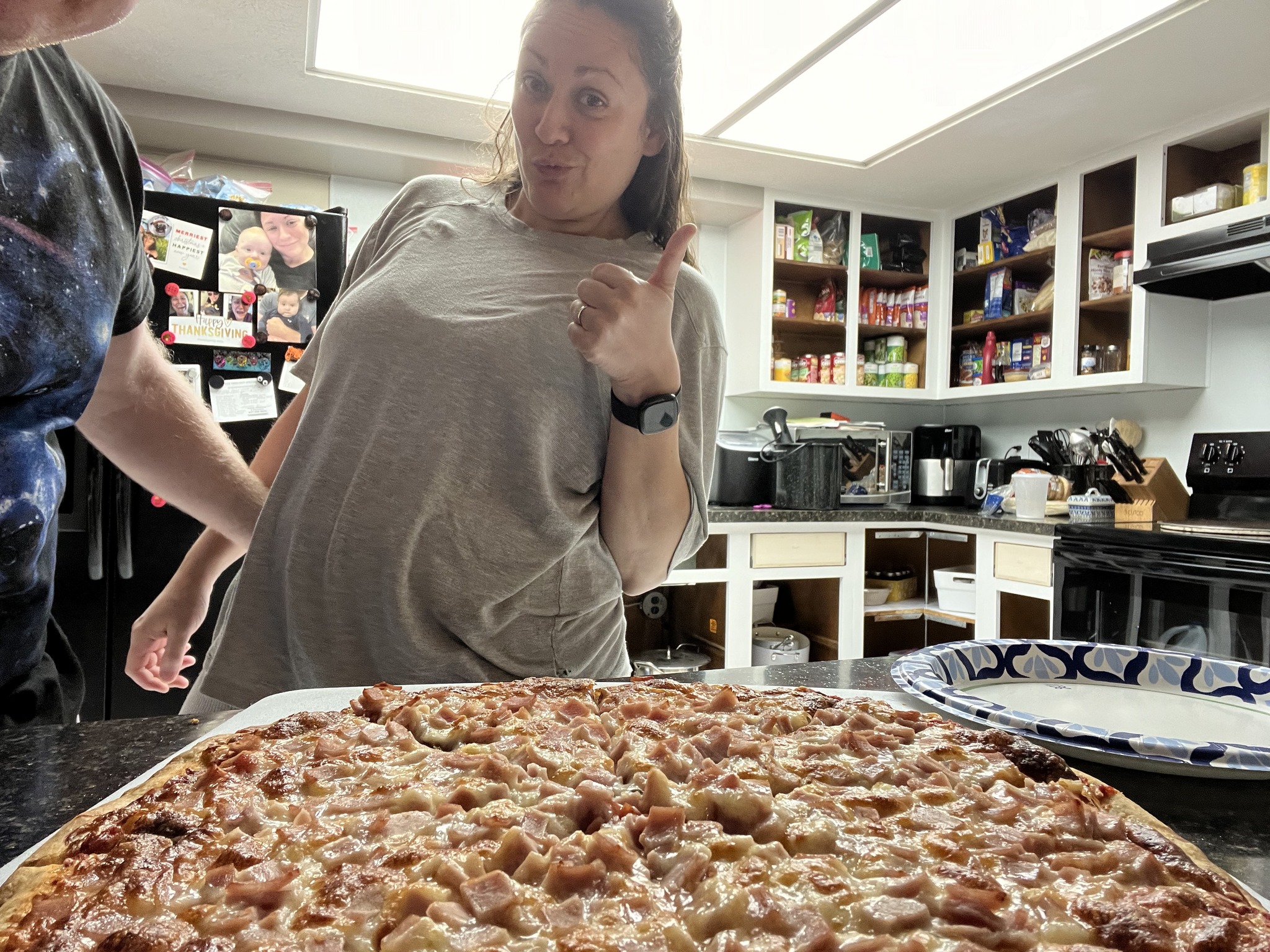 The same homemade pizza in the foreground with a person giving a thumbs-up behind it.