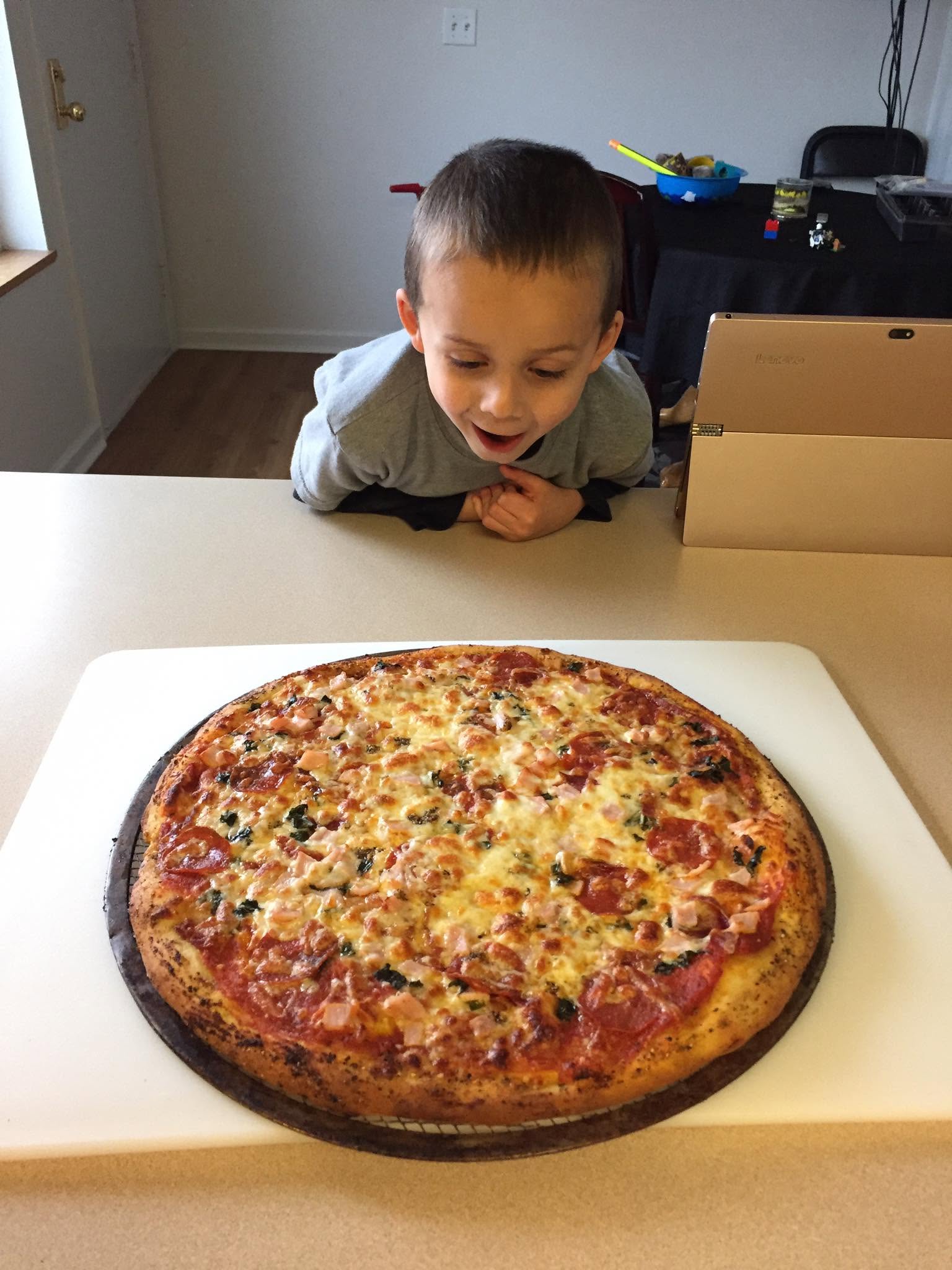 A large baked pizza fills the foreground, with a child looking at it from across the counter.
