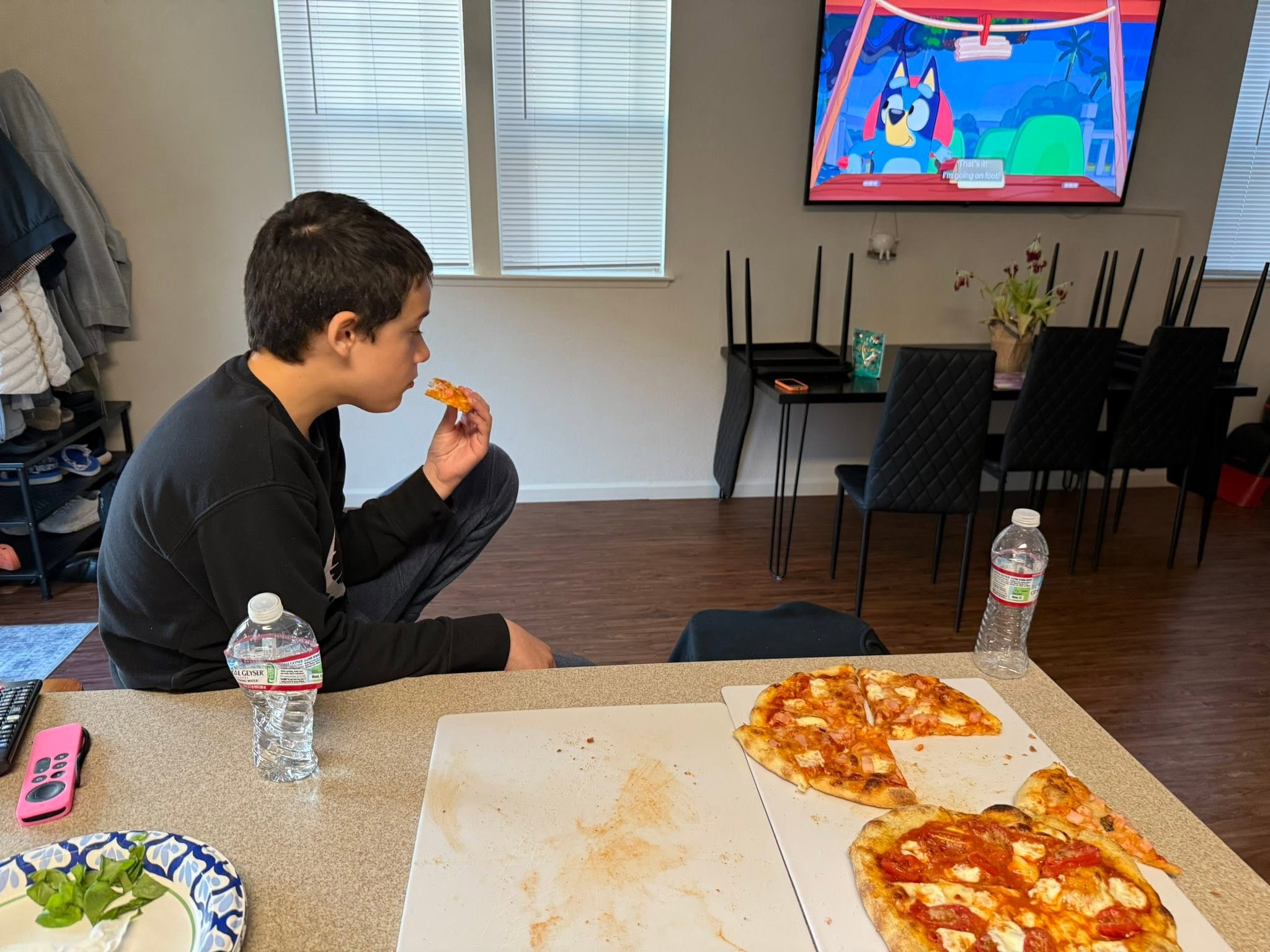 A child eating a pizza slice beside two pizzas while Bluey plays on the television.