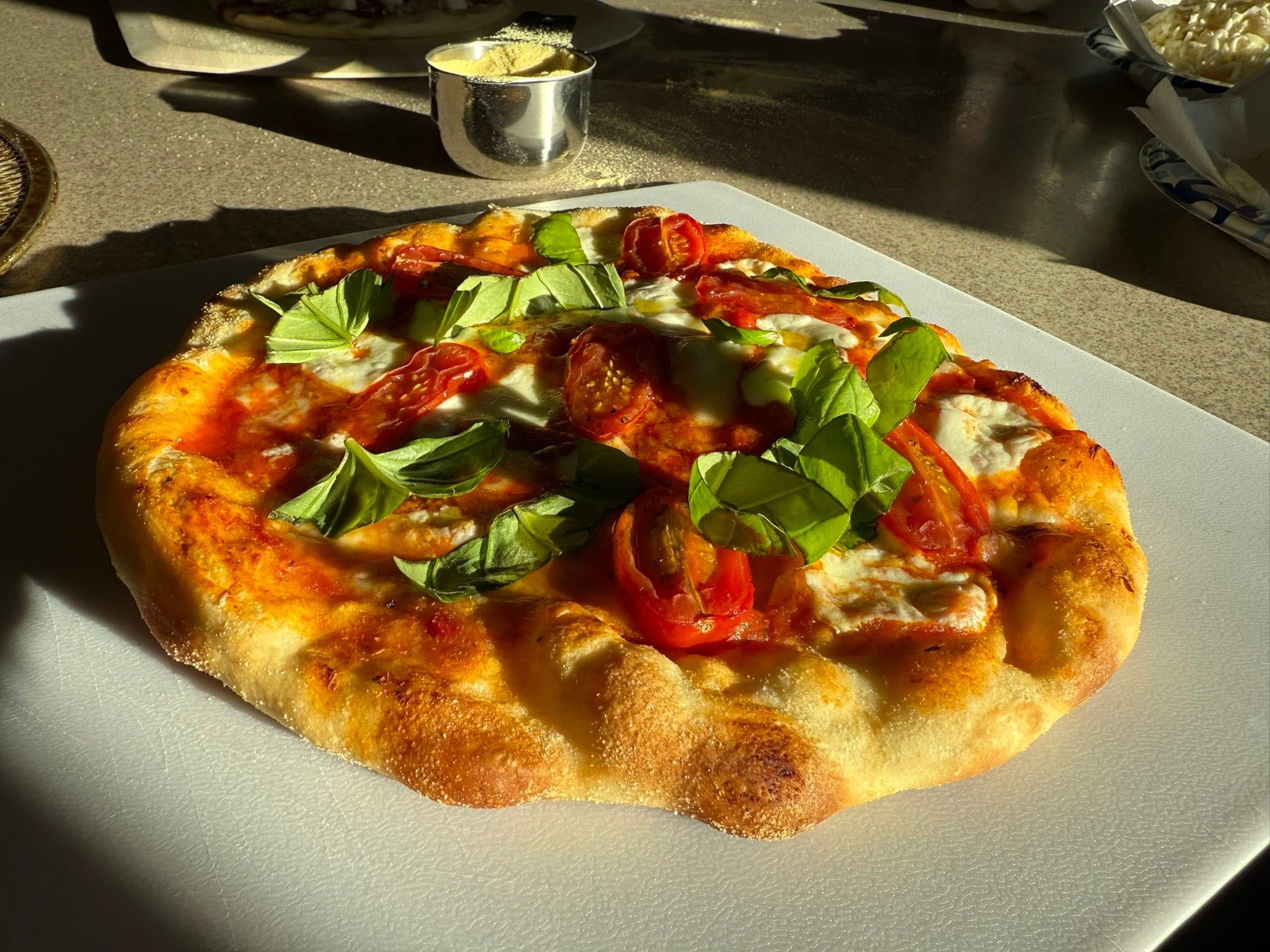 A finished pizza topped with cheese, roasted tomatoes and green leaves, on a board in the kitchen.