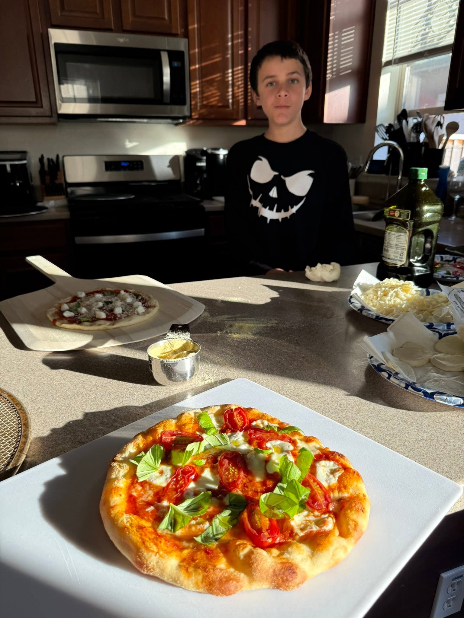 A child at the pizza-making counter with a finished pizza, an uncooked pizza on a peel, and toppings.