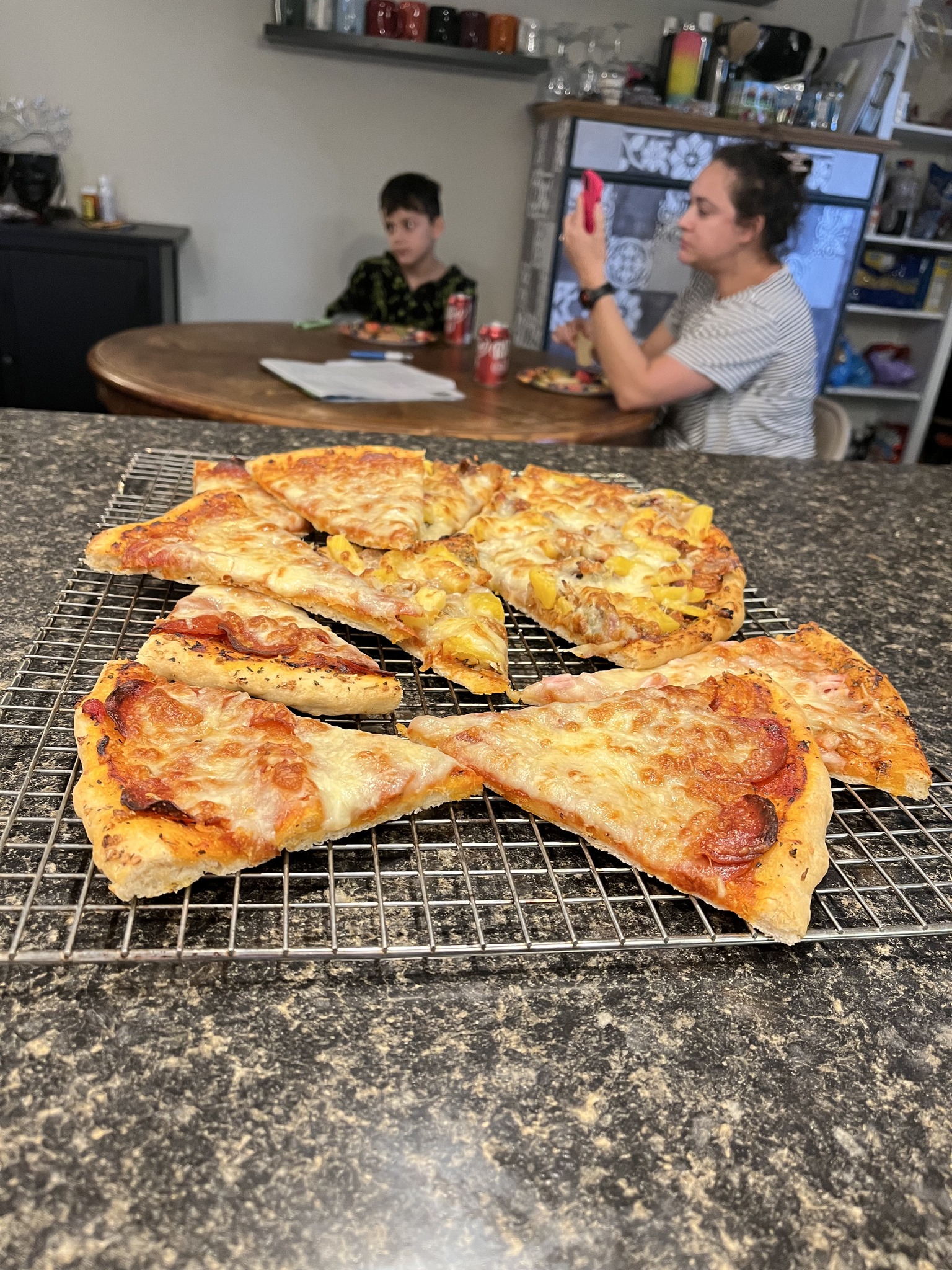 Assorted pizza slices on a rectangular cooling rack with people eating at the table behind them.