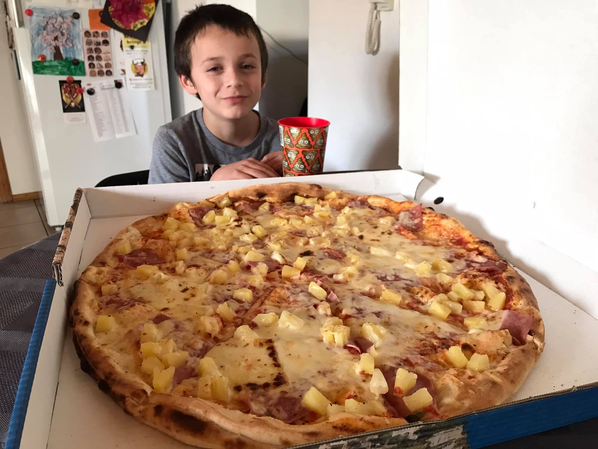 An enormous pizza fills an open takeaway box, with a child sitting behind it for scale.