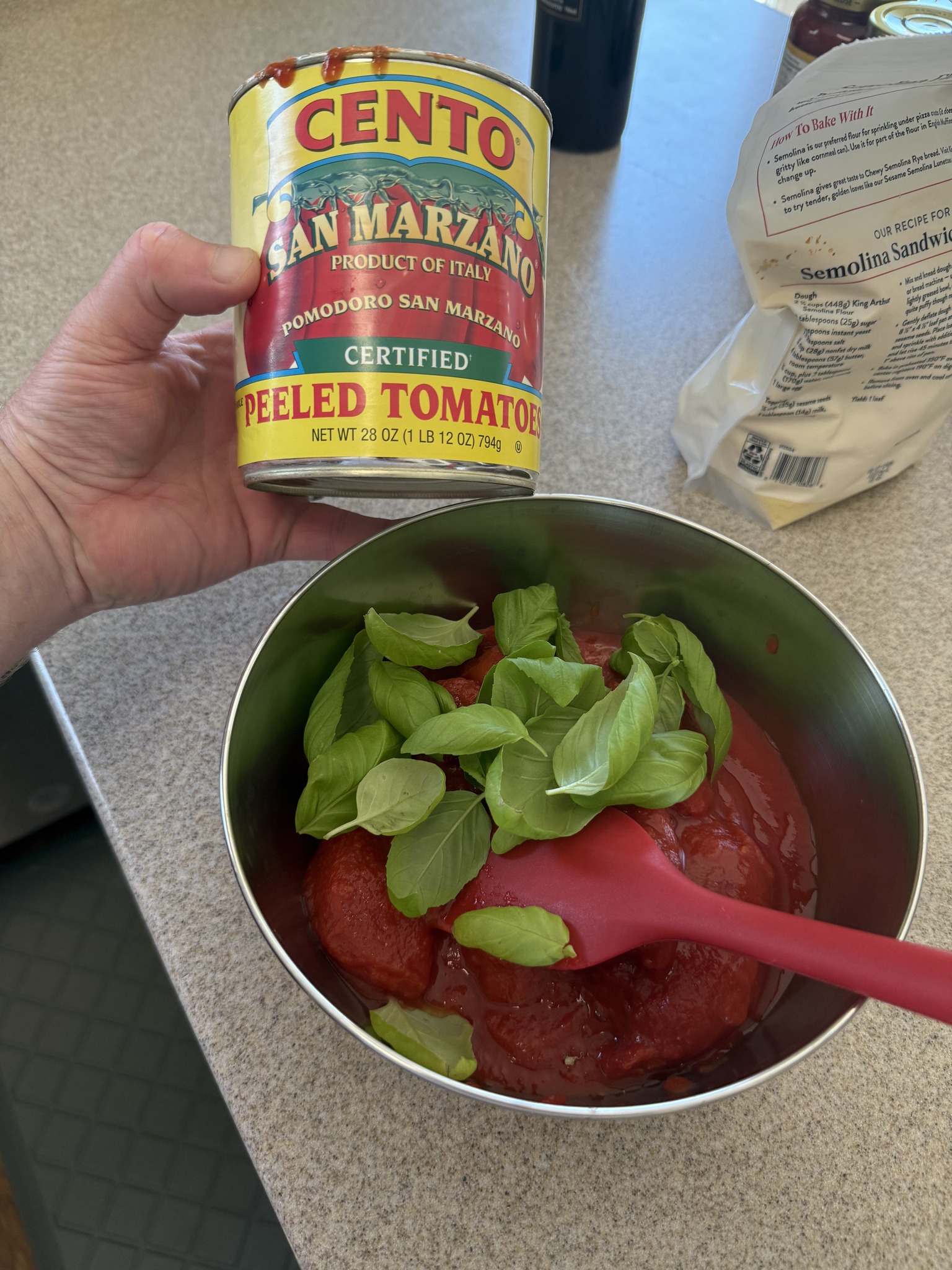 An opened can of San Marzano tomatoes held above a metal bowl of tomatoes and fresh basil leaves.