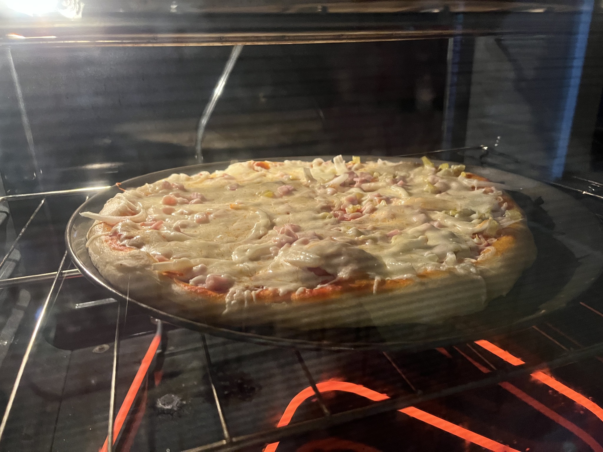 A pizza with onion and yellow-green pieces baking on a round tray inside the kitchen oven.